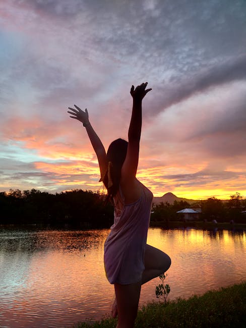 A silhouette of a person standing in a balancing yoga pose outdoors near the water.